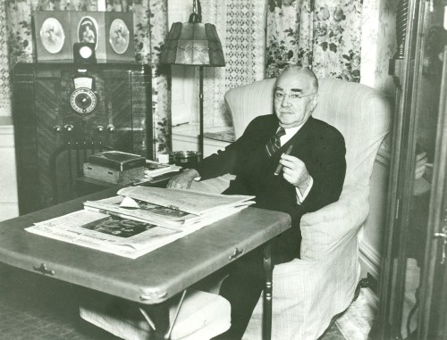 An older man in a suit sits in an armchair, holding a cigar, with a radio and newspapers on a table nearby.