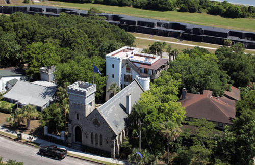 Aerial view of a castle-like building surrounded by trees, with other structures and a road nearby.