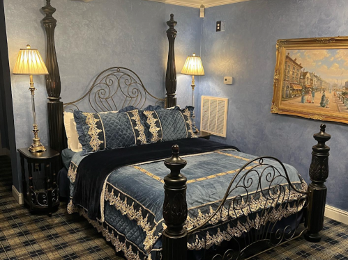 A cozy bedroom featuring a four-poster bed with blue bedding, two lamps, and a framed painting on the wall.