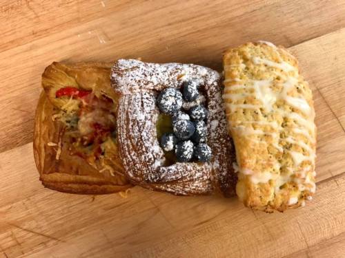 Three pastries on a wooden surface: a savory pastry with cheese, a sweet pastry topped with blueberries and powdered sugar, and a glazed loaf.