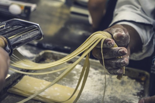 A hand holds freshly made pasta strands, with flour dust on the surface and a pasta machine in the background.