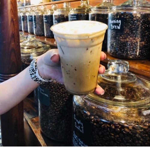 A hand holding a cup of iced coffee in front of jars filled with coffee beans on a wooden shelf.