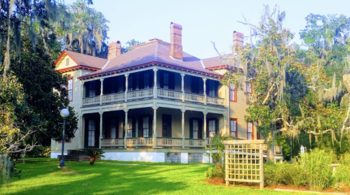 A large, historic house with a wraparound porch, surrounded by lush greenery and trees draped in Spanish moss.