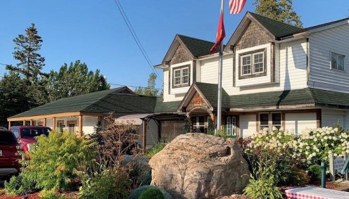 A charming two-story house with a flag, surrounded by lush greenery and colorful flowers in a sunny setting.