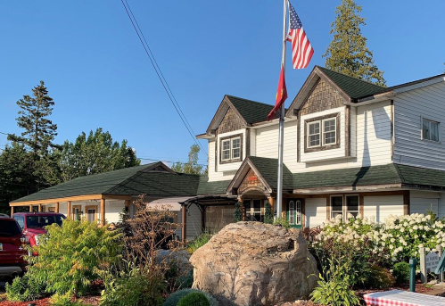 A two-story house with a green roof, American flag, and landscaped garden featuring flowers and a large rock.