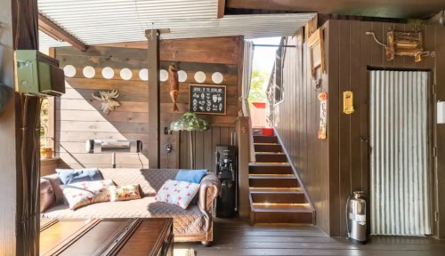 Cozy interior of a wooden cabin featuring a sofa, staircase, and rustic decor. Natural light streams in through windows.