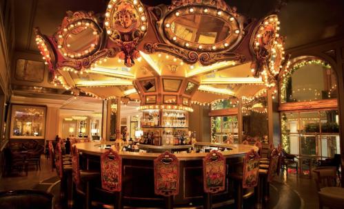 A vintage-style bar with ornate mirrors and warm lighting, featuring a circular counter and decorative chairs.