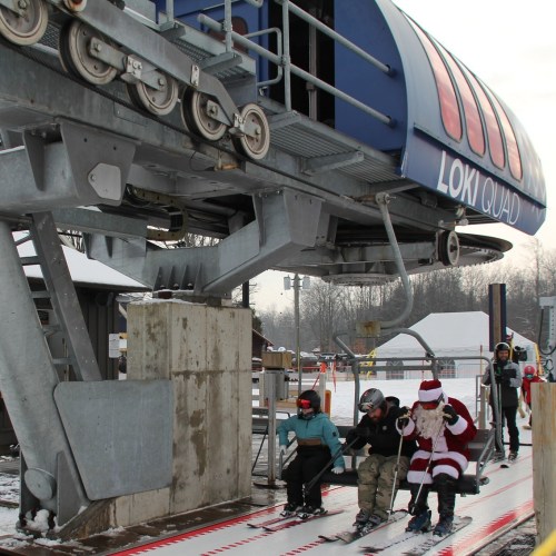 A ski lift named "Loki Quad" with skiers boarding, including a person in a Santa costume. Snowy winter scene.