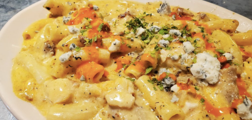Creamy pasta dish topped with herbs, blue cheese, and a drizzle of spicy sauce on a light pink plate.