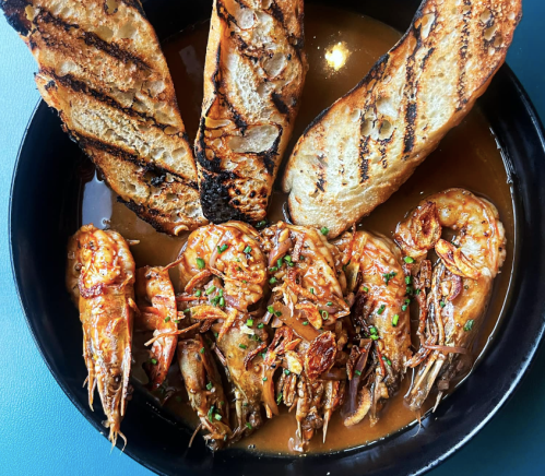 A bowl of shrimp in a savory sauce, garnished with crispy fried onions, accompanied by grilled bread slices.