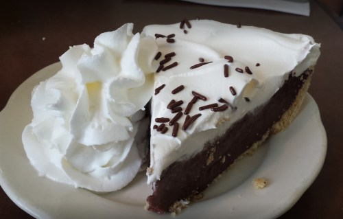 A slice of chocolate cream pie topped with whipped cream and chocolate sprinkles on a white plate.