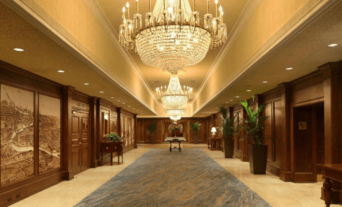 Elegant hallway with chandeliers, wooden paneling, and decorative wall art, featuring a plush carpet and potted plants.
