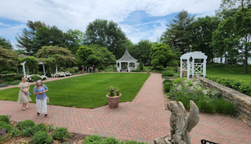 A lush garden with a gazebo, surrounded by greenery and flowers, featuring people walking along a brick path.