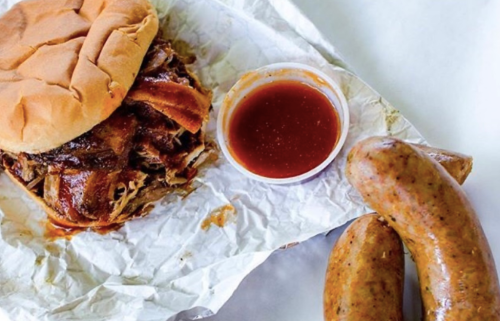 A pulled pork sandwich with barbecue sauce, accompanied by a side of sausage and a small cup of sauce.