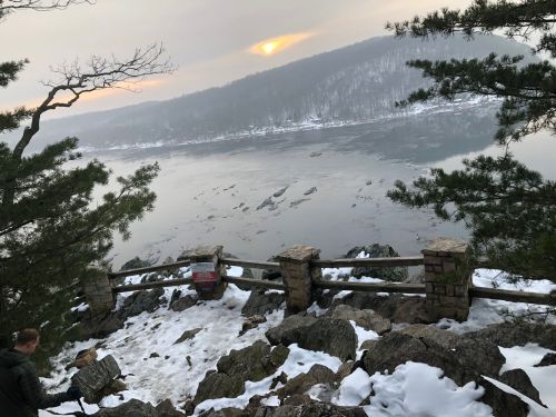 A serene winter landscape with a river, rocky shore, and a sunset partially obscured by misty mountains.