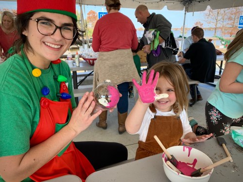 A smiling woman in a festive outfit holds a decorated ornament, while a girl shows her pink-painted hand at a craft event.
