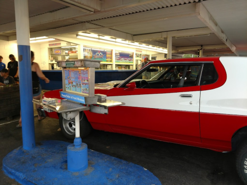 A red and white vintage car parked at a drive-in with a menu stand nearby and people in the background.