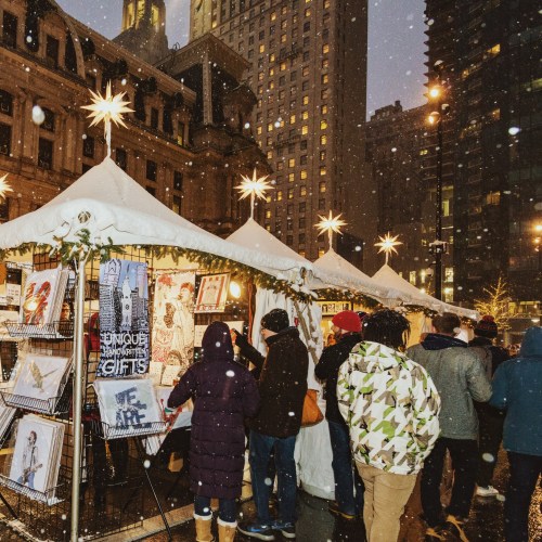A festive market scene with people browsing art and gifts under snow, surrounded by city buildings and holiday lights.