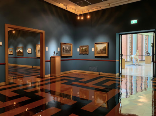 A gallery interior featuring framed paintings, polished floors, and a doorway leading to another room with a sculpture.