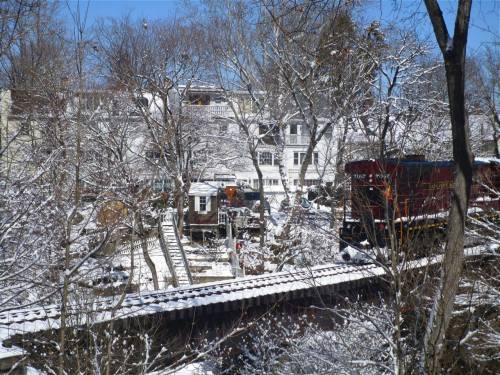 A snowy landscape featuring a train track, a red train, and houses surrounded by trees in winter.