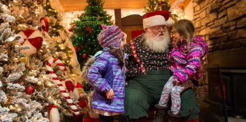 Santa Claus sits in a cozy, decorated room with two children, surrounded by Christmas trees and festive decorations.