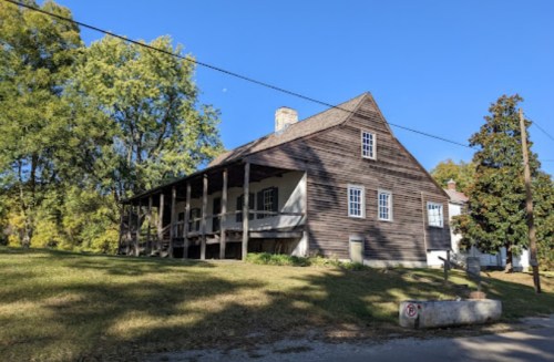 Historic wooden house with a porch, surrounded by trees and a clear blue sky.