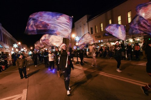 A parade at night with participants waving colorful flags, festive lights, and a crowd watching along the street.