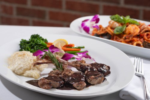 A plate of lamb chops with a rich sauce, served with vegetables and creamy risotto, alongside a pasta dish.