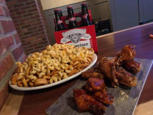 A plate of macaroni and cheese with fries, alongside a platter of chicken wings and a six-pack of beer.