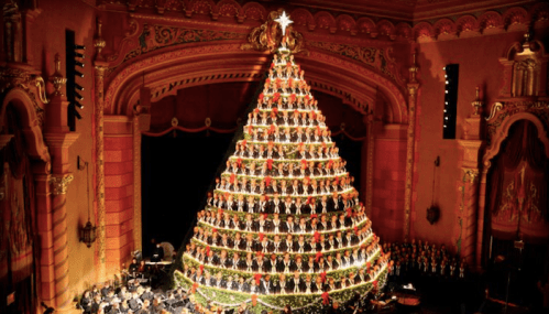 A large, decorated Christmas tree made of lights and ornaments, set on a stage with musicians and a choir in the background.