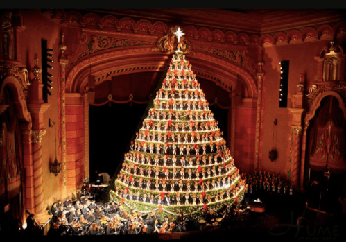 A large, decorated Christmas tree made of lights and ornaments, set on a stage with musicians in the background.
