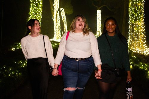 Three women walk hand in hand through a lit pathway, smiling and enjoying a festive atmosphere.