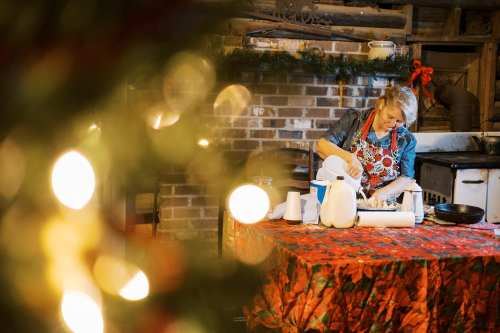 A woman prepares food in a rustic kitchen, decorated for the holidays, with a festive tablecloth and Christmas lights in the foreground.