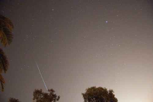 A night sky filled with stars, featuring a bright meteor streaking across the scene above silhouetted trees.