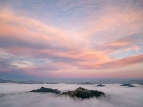 A serene landscape of rolling hills above a sea of clouds, with a colorful sunset sky filled with soft pink and blue hues.