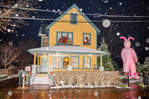 A brightly colored house decorated for the holidays, with a large pink inflatable figure and snow falling.