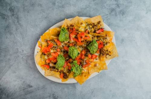 A plate of nachos topped with ground beef, melted cheese, diced tomatoes, and green salsa on a gray background.