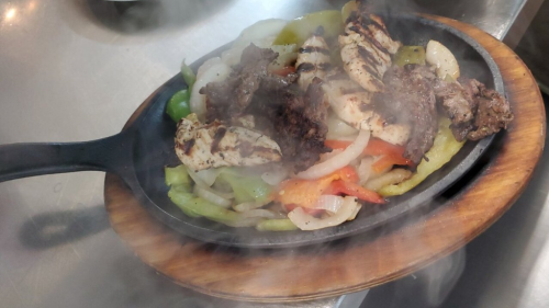 Sizzling fajitas on a wooden platter, featuring grilled chicken, beef, and colorful bell peppers, with steam rising.