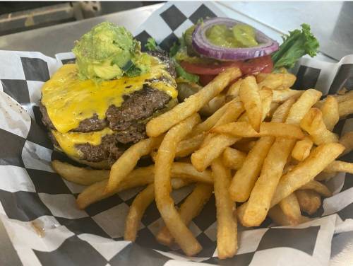 A cheeseburger topped with avocado, served with a side of golden French fries and fresh toppings on a checkered plate.