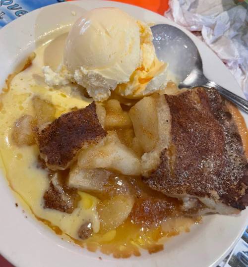 A bowl of warm apple cobbler topped with vanilla ice cream and drizzled with cream.