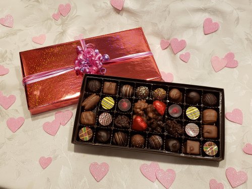 A box of assorted chocolates with a shiny red gift box, surrounded by pink heart decorations on a light tablecloth.