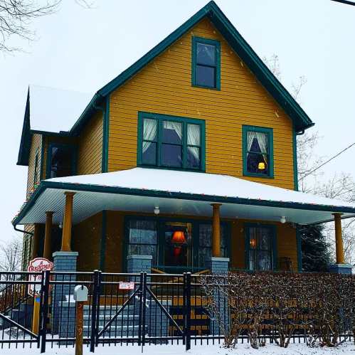 A yellow house with a snow-covered roof, surrounded by a fence, in a winter landscape. Snowflakes are falling.