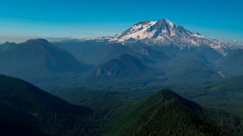 A panoramic view of a snow-capped mountain surrounded by lush green forests and rolling hills under a clear blue sky.
