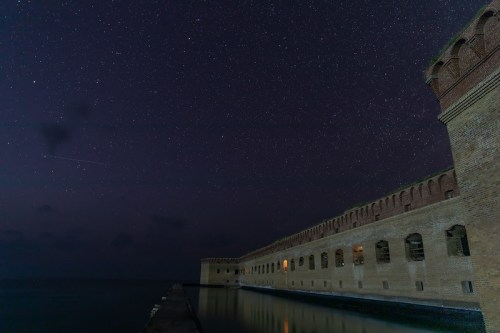 A historic brick structure by the water under a starry night sky, reflecting in the calm surface below.