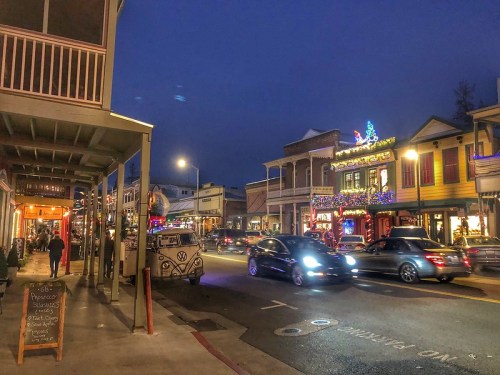 A lively street scene at dusk, featuring shops, cars, and festive lights adorning buildings.