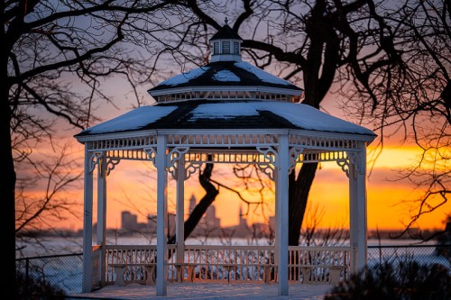 A snow-covered gazebo silhouetted against a vibrant sunset, with city skyline in the background.