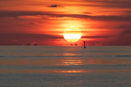 A vibrant sunset over calm waters, with a silhouette of a marker buoy in the foreground against a colorful sky.