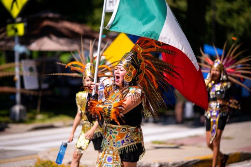 A woman in vibrant traditional attire waves a Mexican flag while participating in a lively parade.