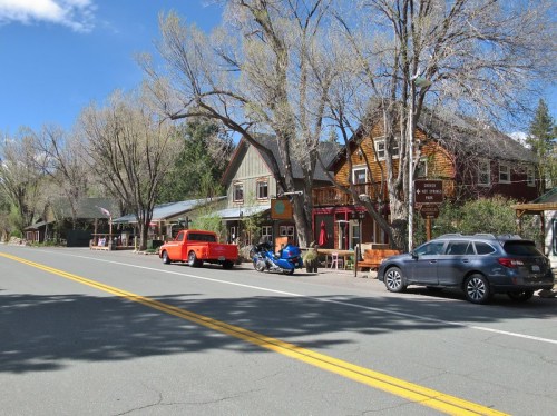 A quiet street lined with rustic buildings, parked cars, and trees under a clear blue sky.