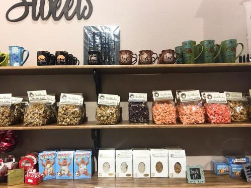 A shelf displaying various popcorn flavors, mugs, and gift items in a cozy shop setting.